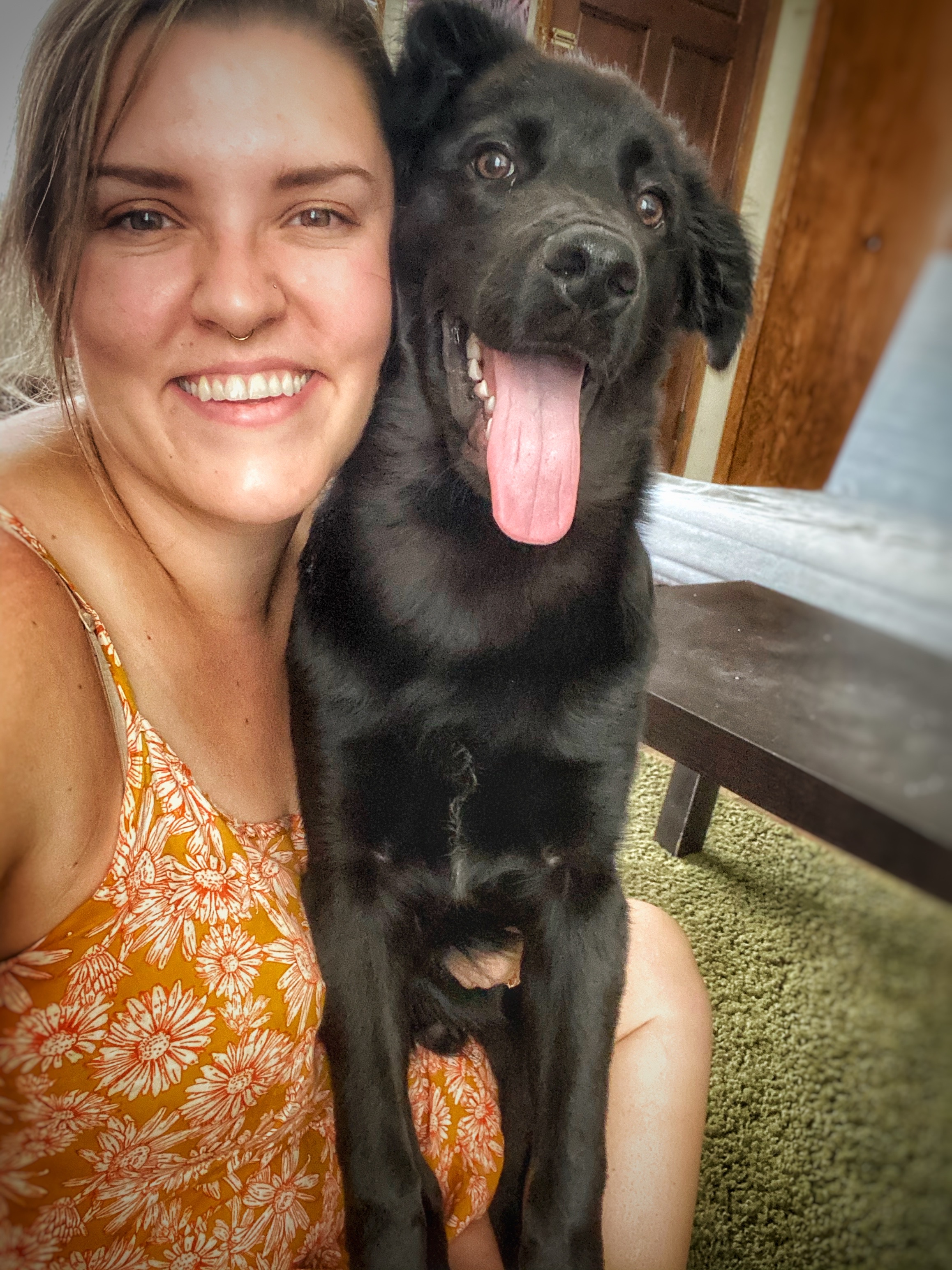 Chef Heather sitting in a yellow dress with daisy print and with black dog (Brewer) smiling at camera
